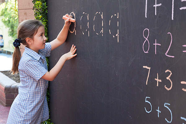 A young girl in school uniform writes words on a chalk board